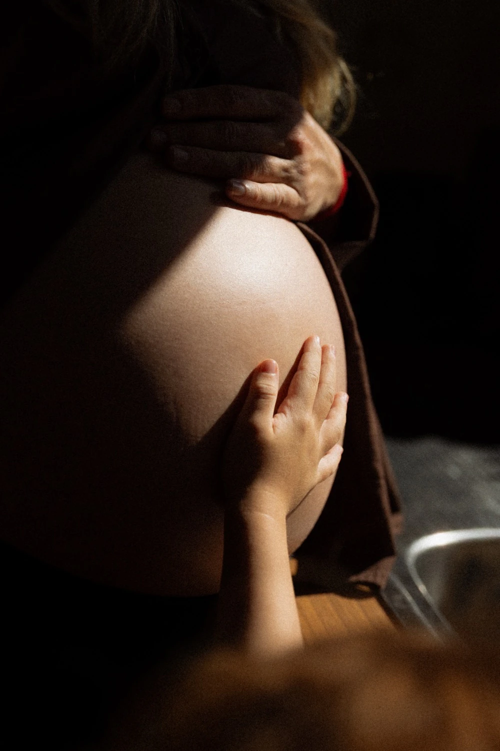 A pregnant woman's belly lit from one side, with an adult's hand resting above it and a child's hands below.