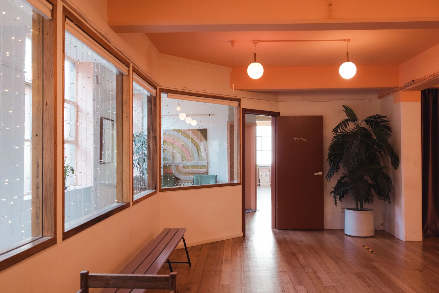 The entry at Studio Take Care: a bench under a run of windows strung with fairy lights, a potted palm in the corner, and an open door through to a bright room beyond.