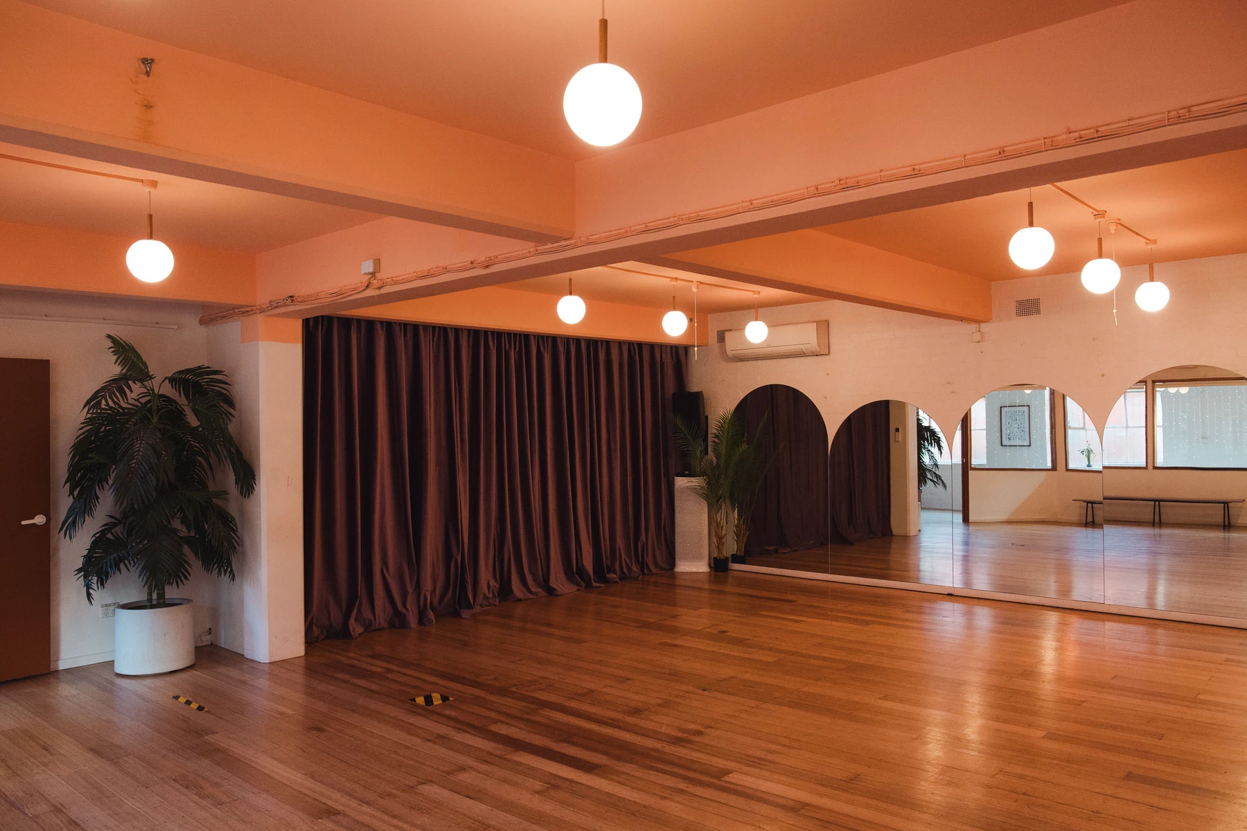 The main room at Studio Take Care: polished timber floors under a wall of full-height mirrors, a deep red curtain drawn across the wall beside them, a potted palm by the door and round pendant lights overhead.
