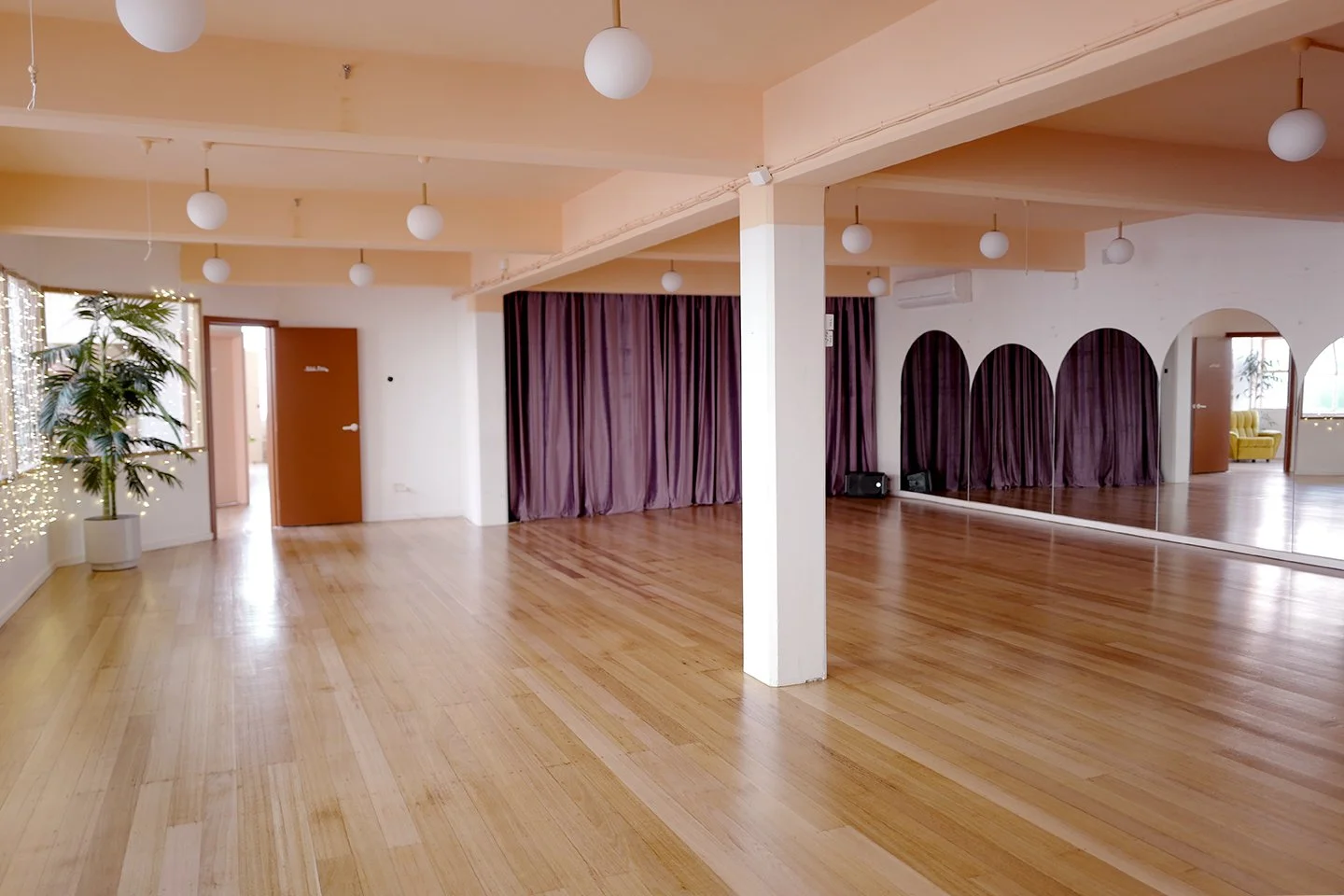 The main room at Studio Take Care: a wide, empty studio with pale timber floors, peach walls, round pendant lights and full-height mirrors along one side.